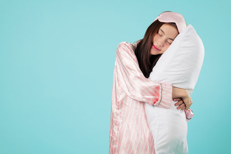 Happy asian woman in pajamas, sleep mask, hugs a pillow, smiling after a good night sleep, feeling rested, embracing the pillow with closed eyes in morning mood, headshots isolated blue backgroundの写真素材