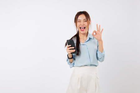 Happy excited young Asian woman smile holding a mobile phone and showing an ok gesture sign. She is very happy with her smart phone. isolated studio white backgroundの写真素材