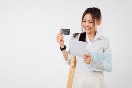 Smiling Asian woman accident with broken arm bone in plaster cast holds bills with credit card,...