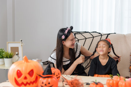 A woman braids a childs hair, both are in costume for a happy halloween day, small pumpkins decorate the scene near a spider web on the sofa.の写真素材