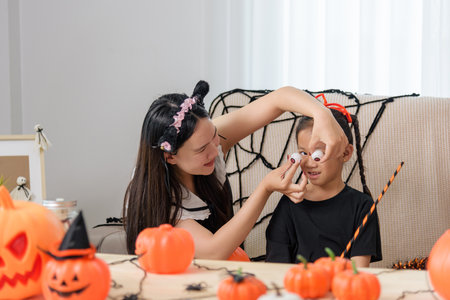 A woman with cat ears prepares a girl for halloween, the woman holds fake eyeballs near the childs face as orange pumpkin decorations sit on the table near a spooky spider web.の写真素材