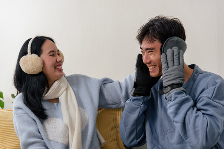 Happy couple enjoys cozy home on winter day people laughing indoors wearing warm clothing like fluffy earmuffs and protective gloves for chilly season creating comfortable winter atmosphere.の写真素材