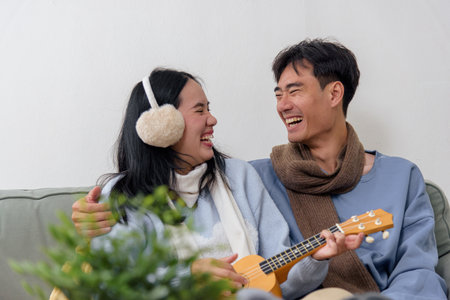 A cheerful couple celebrates winter holidays, the woman wearing earmuffs laughs, while the man wearing a scarf plays the ukulele with joy, sharing a happy moment together on the sofa.の写真素材