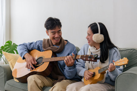 Winter couple enjoys happy indoor home time, people playing music on guitar and ukulele, smiling and laughing together creating cozy atmosphere on cold winter day inside apartment.の写真素材