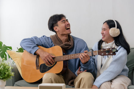 Winter couple happy people enjoy cozy home indoor moments, man plays acoustic guitar for woman, both laughing with joy, celebrating cold winter day with warm peaceful comfort.の写真素材