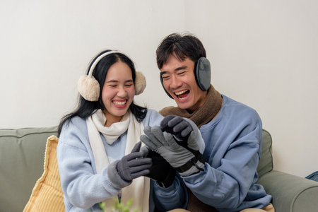 Winter day brings happy couple home, people enjoying cozy indoor moment on sofa, sharing laughter, wearing warm sweaters, scarves, earmuffs, gloves for chilly winter season, feeling joyful together.の写真素材
