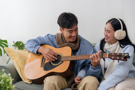 Winter day finds happy couple at home, young people enjoying relaxing indoor comfort, man playing acoustic guitar for smiling woman, warm sweaters cozy scarf comfort chilly winter day.の写真素材