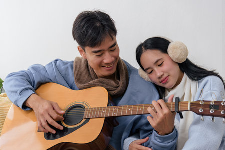 Winter day indoors, happy young couple enjoys cozy home bonding with music, man plays guitar, woman rests on shoulder, creating warm winter memories.の写真素材
