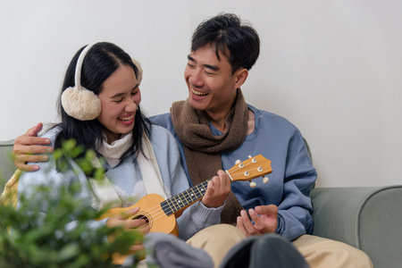 Winter couple shares happy home indoor time, people enjoying cozy moments playing ukulele, man wearing scarf and woman earmuffs for warm day, creating joyful winter memories.の写真素材