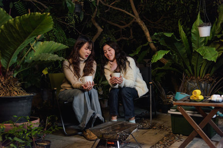 Winter night, women friends happy camp outdoor forest people enjoying warm drinks by glowing grill, creating cozy winter ambiance, celebrating friendship in lush foliage.の写真素材