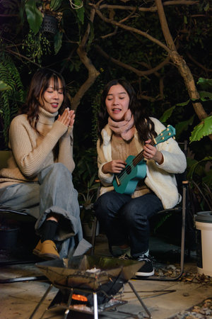 Winter, happy women camp outdoor with friend, people enjoy forest night, companion plays ukulele, other claps, cozy winter evening fun.の写真素材