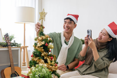 Christmas couple share love, happy holiday winter celebration, decorating home christmas tree, woman capturing cheerful moment on phone, man placing star topper, creating festive atmosphere.の写真素材
