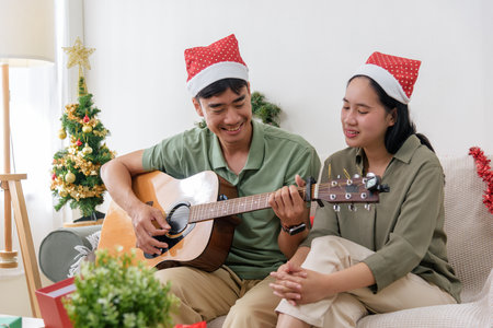 Christmas couple shares love, happy holiday celebration, Santa hats on, man plays guitar, woman enjoys music, winter day brings joy, cozy Christmas gathering.の写真素材