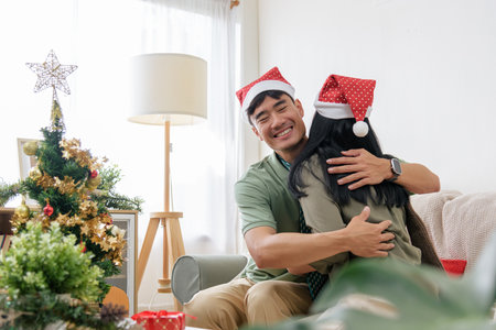 Christmas happy couple embraces, showing love in cozy winter home, wearing santa hats, celebrating christmas holiday with decorated tree and gift, warm festive atmosphere.の写真素材