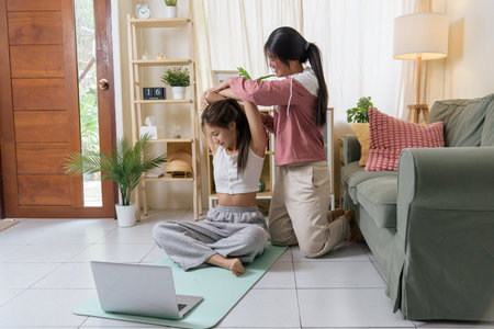 Young women workout yoga active teen exercising sport friend stretching arms, receiving helpful guidance from partner, enjoying home fitness training with online instruction via laptop on floor.の写真素材