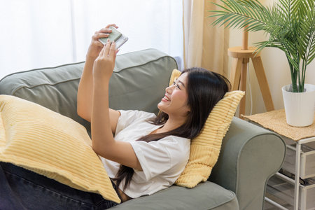 Happy young woman using mobile phone, person enjoys cell smartphon for online browsing, relaxing comfortably on soft sofa in bright home living room, enjoying quiet moment indoors.の写真素材
