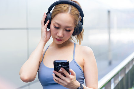 Fit young sport woman prepares for outdoor workout checking phone, selecting energizing music using headphones, ready for active fitness exercise, healthy urban lifestyle workout.の写真素材
