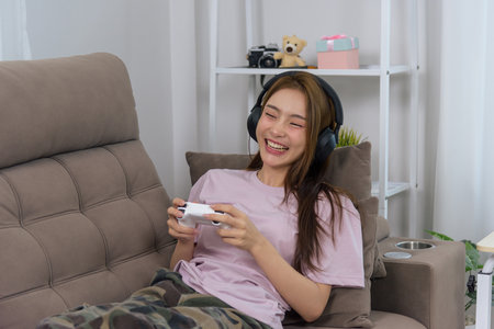 Happy young woman at home, people find her enjoying activity playing video games, wearing headphones, laughing, relaxing on sofa for leisure and entertainment.の写真素材