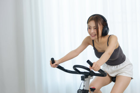 Young woman, happy people, enjoys home fitness activity, cycling on stationary bike with wireless headphones, radiating joy during workout.の写真素材