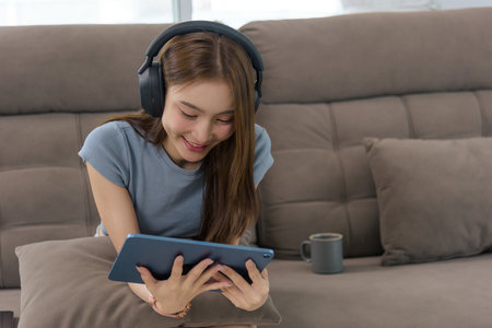 Young woman at home enjoys happy activity, people relaxing, wearing headphones while watching digital tablet, discovering media content on comfortable couch, embracing modern leisure time.の写真素材