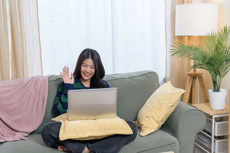 Happy teen woman relaxing at home, uses laptop for virtual meeting, waving hand, comfortable sofa...