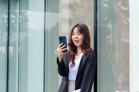 Young Asian business woman in suit on street, busy with phone communication for work, leaning...
