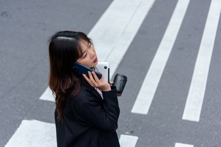 Young Asian business woman in suit works on street, busy with phone call, carrying laptop, travel mug, depicting daily work commute in vibrant urban city environment, successful professional life.の写真素材