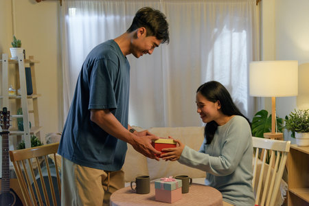 Happy young couple in love, man gives woman a special red gift box for romantic valentine day celebration at home, sharing affection and joy indoors with lovely atmosphere.の写真素材