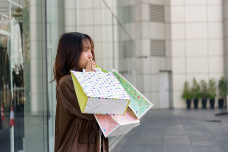 Young Asian woman feeling joy from successful shopping, proudly holding multiple colorful fashion bags after shop visit, posing outside modern city mall, representing people enjoying lifestyle.の写真素材