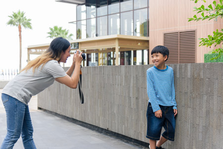 Mother, child, son enjoy summer city street, woman photographs happy family outing, parent captures smiling boy standing against urban wall outdoors, warm summer day.の写真素材