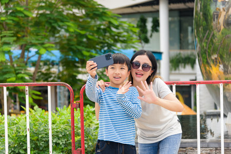 Mother and child, family woman, parent, son enjoy summer city street selfie, happily waving for camera, outdoor urban street scene with people around.の写真素材