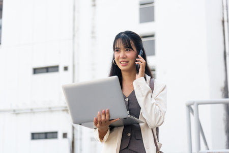 Smiling happy Asian woman for business work outdoor on street, talking on mobile phone, using laptop, representing modern urban city lifestyle for young professionals.の写真素材
