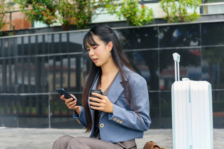 Travel woman, business person outdoor lifestyle on city urban street, engages work using phone, enjoys coffee moment, casual scene for people, luggage nearby representing journey.の写真素材