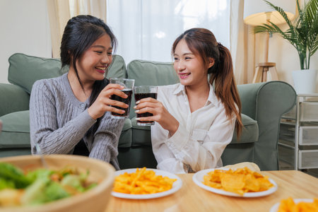 Young women enjoy eat snacks, friend smiling, clinking drinks, people relax in cozy living room with tasty food and refreshing drinks, sharing joyful moments at home.の写真素材