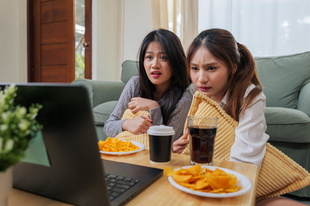Two young women friends enjoy watching laptop entertainment, eat crunchy potato chips, sip refreshing drinks, showing engaged expressions, focused on screen during casual leisure living room home.の写真素材