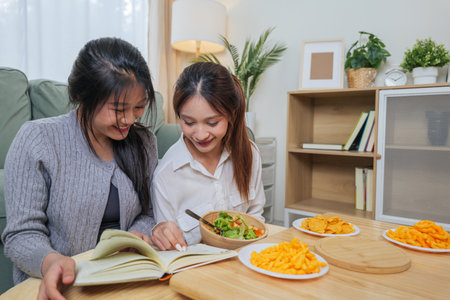 Two young women, close friends, truly enjoy eating delicious crunchy snacks and refreshing healthy salad, sharing interesting book together in warm, inviting living room setting for relaxation.の写真素材