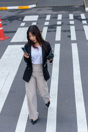 Young Asian woman, business professional, walks busy urban street crosswalk, looking at smartphone,...