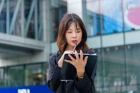 Young woman, business suit, work on street, busy planning tasks, holding notebook and pen, showing professional dedication, contemplating future ideas in vibrant urban setting.の写真素材