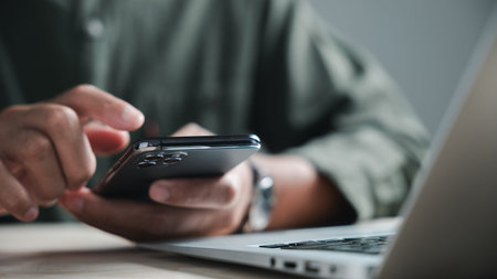 Close-up of a person's hand holding a smartphone with a laptop in the background. The image focuses...