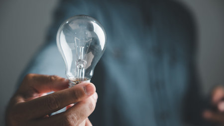A person's hand holding a clear light bulb against a blurred blue background. The bulb is empty and...