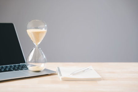 Hourglass time management concept shows sandglass timer with sand flowing near laptop and notebook...