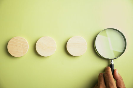 Three wooden circles arranged in a row, with a magnifying glass positioned to the right.  A hand holds the magnifying glass.  The background is a light green.の写真素材