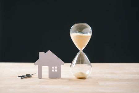 Hourglass with flowing sand, small model house, and keys symbolize real estate time constraint,...