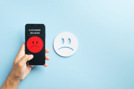 Survey shows customer online review mobile phone, hand holds smartphone with red unhappy feedback, poor service rating low satisfaction experience, white sad face visible.の写真素材