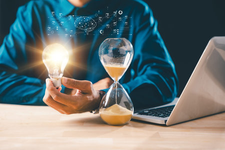 Businessman holding bright lightbulb, a bulb of innovation idea, beside hourglass with flowing sand, powerful concept showing business time management, countdown, and deadline importance.の写真素材