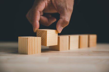 A hand placing a wooden block on top of other wooden blocks. The blocks are arranged in a row on a...