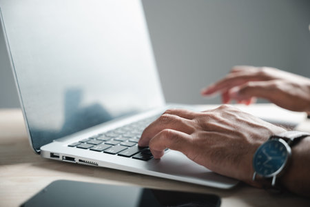 Close-up of a hand typing on a laptop keyboard. A wristwatch with a blue face is visible.