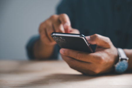 Close-up of a person's hands holding a smartphone, with a wristwatch and wooden table in the...