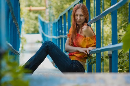 Young woman with red hair and yellow jacket sitting on a small bridge and posing in summer day, telephotoの写真素材