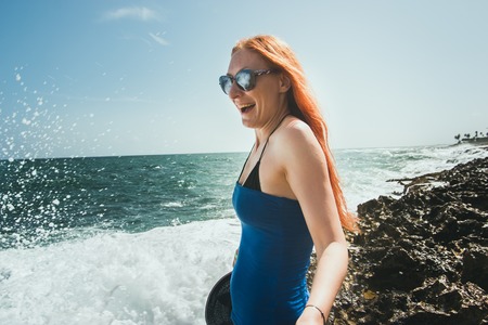 Young woman with red hair and sun glasses standing on sea coast and playing with water spray, close upの写真素材